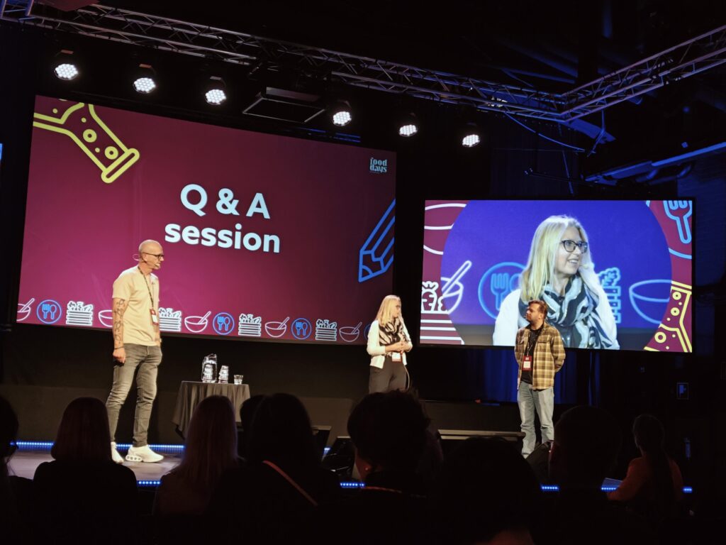 Women and two men speakers are standing on the stage and answering questions from the audience. Behind them there is a big screen with the "Q&A" text and on the othyer screen there is a closer look on the woman speaker.