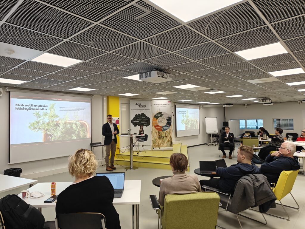 Lignell & Piispasen Kehitysjohtaja Harri Nylund esittää luokkatilan edustalla. Taustalla näkyy esitys ja Agri-Food klutserin rollupit. Edustalla näkyy osallistujia.