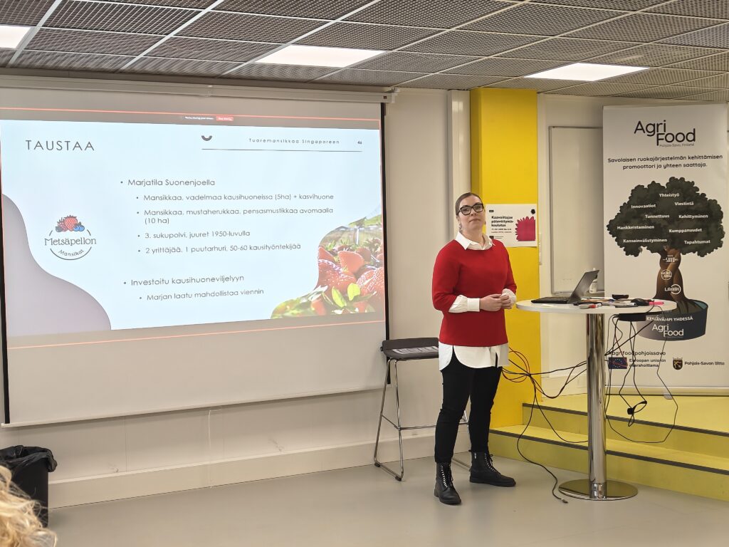 Metsäpellon mansikkatilan yrittäjä Pauliina Kovanen esittää luokkatilan edustalla. Taustalla näkyy esitys ja Agri-Food klutserin rollupit.