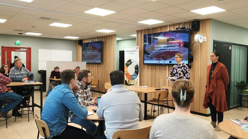 Ryhmä ihmisiä osallistuu työpajamaiseen tilaisuuteen modernissa tilassa. Osallistujat istuvat pöytien äärellä ja kaksi henkilöä seisoo esityksen edessä. Seinällä näkyy suuri näyttö, jossa on tapahtuman dia ja teksti 'Elinvoimaa paikallisilla elintarvikehankinnoilla."